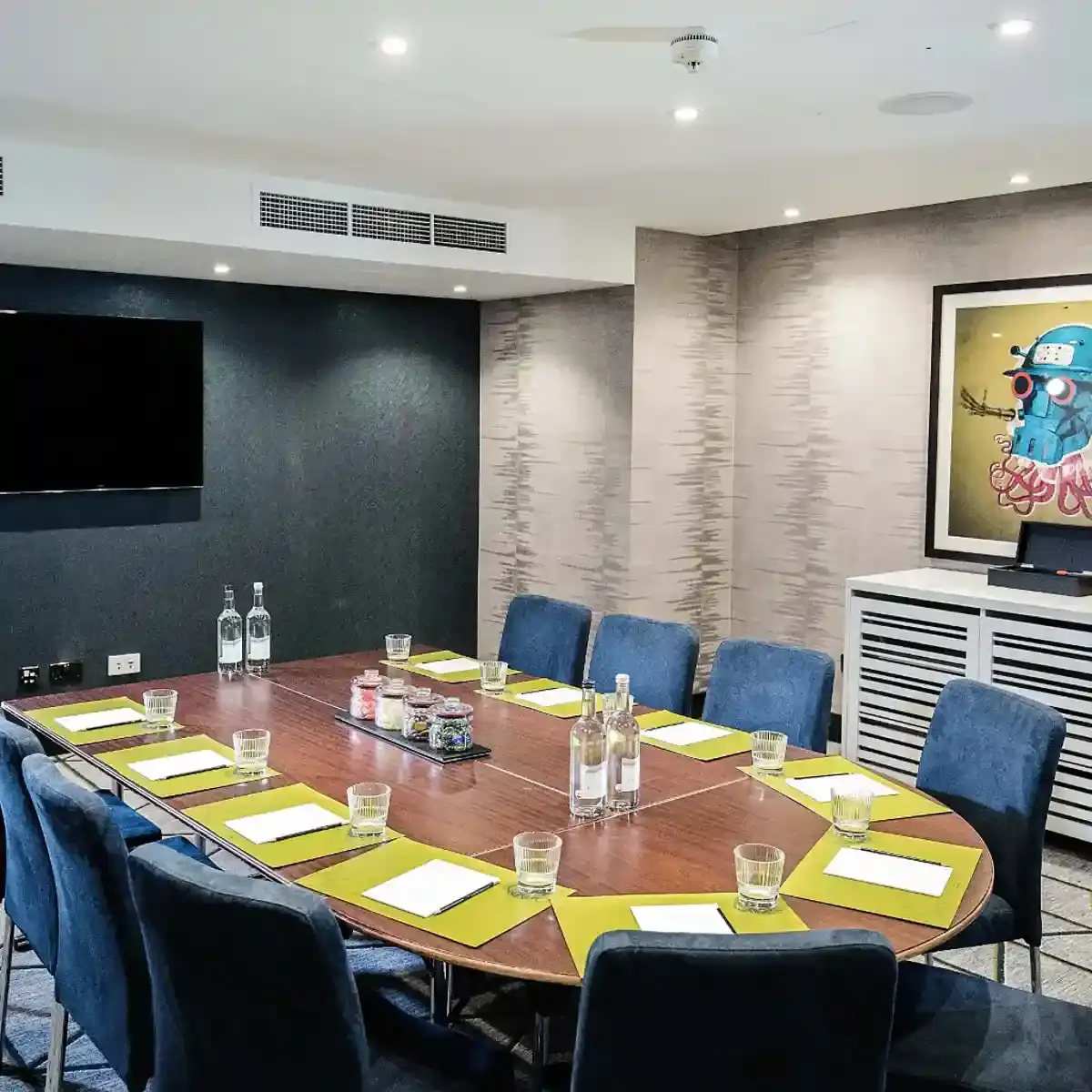 A semi oval meeting table with blue fabric seating around the edges. Several lime green placemats can be seen each with a writing pad and pencil and drinking glass.