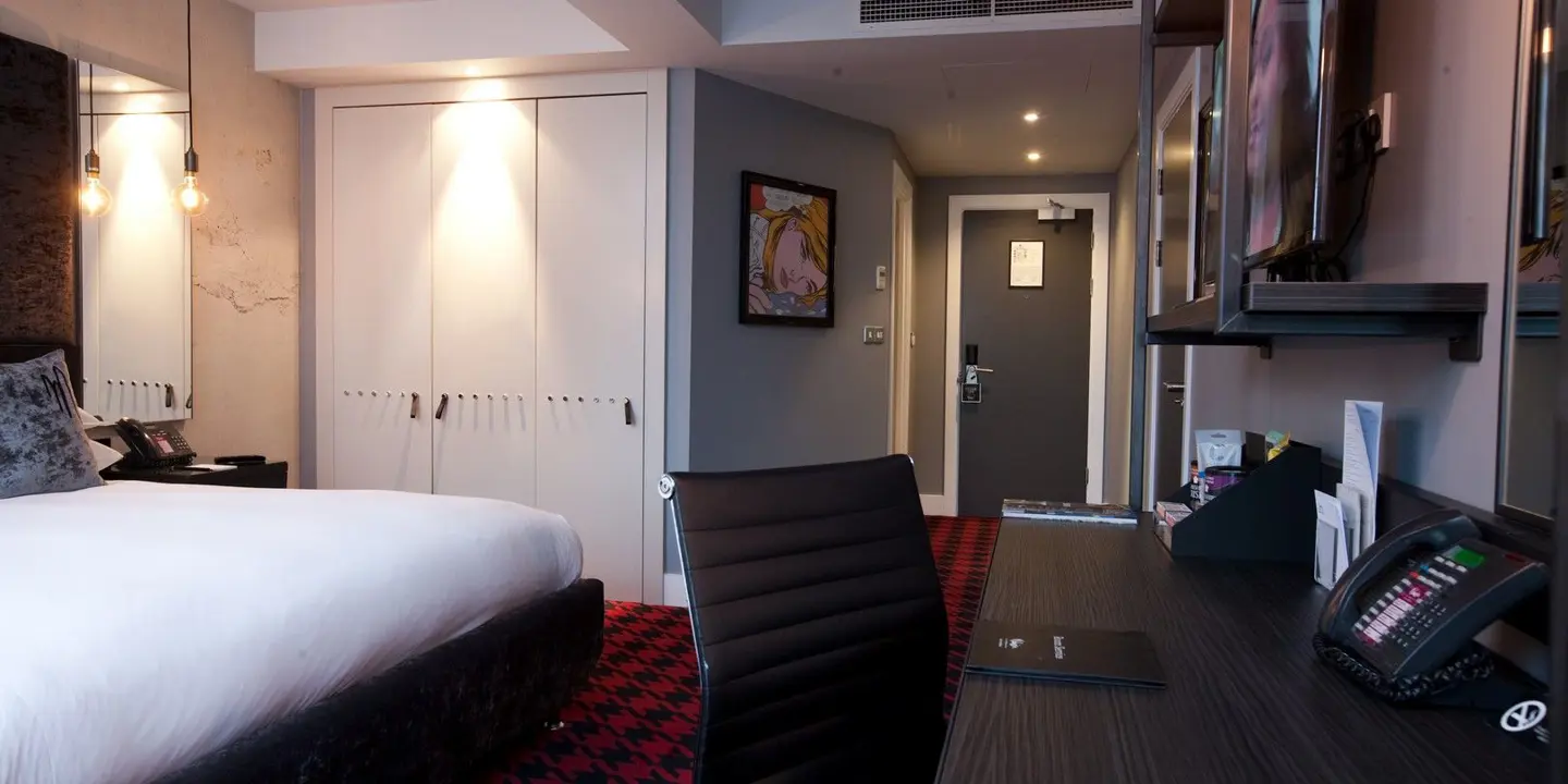 View of desk and chair and standard double bed with white linen sheets a red houndstooth carpet and flat screen TV