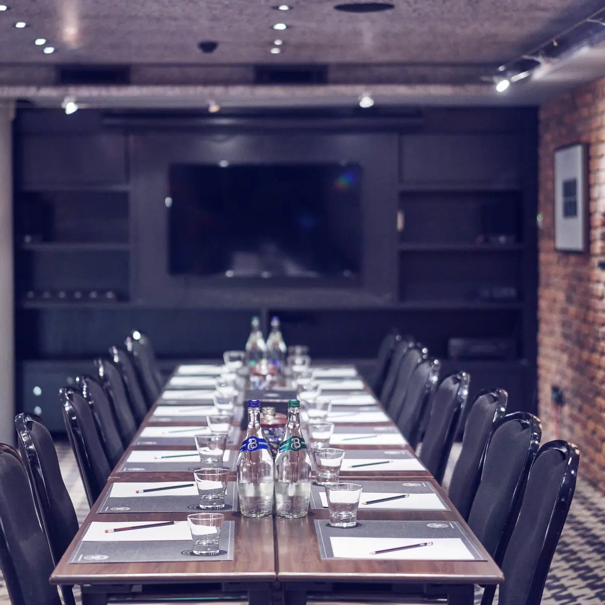 View of a meeting table with 16 black chairs on a black and white carpet, an exposed brick wall with artwork is to the right of the table, and a flat screen TV can be seen in the background.