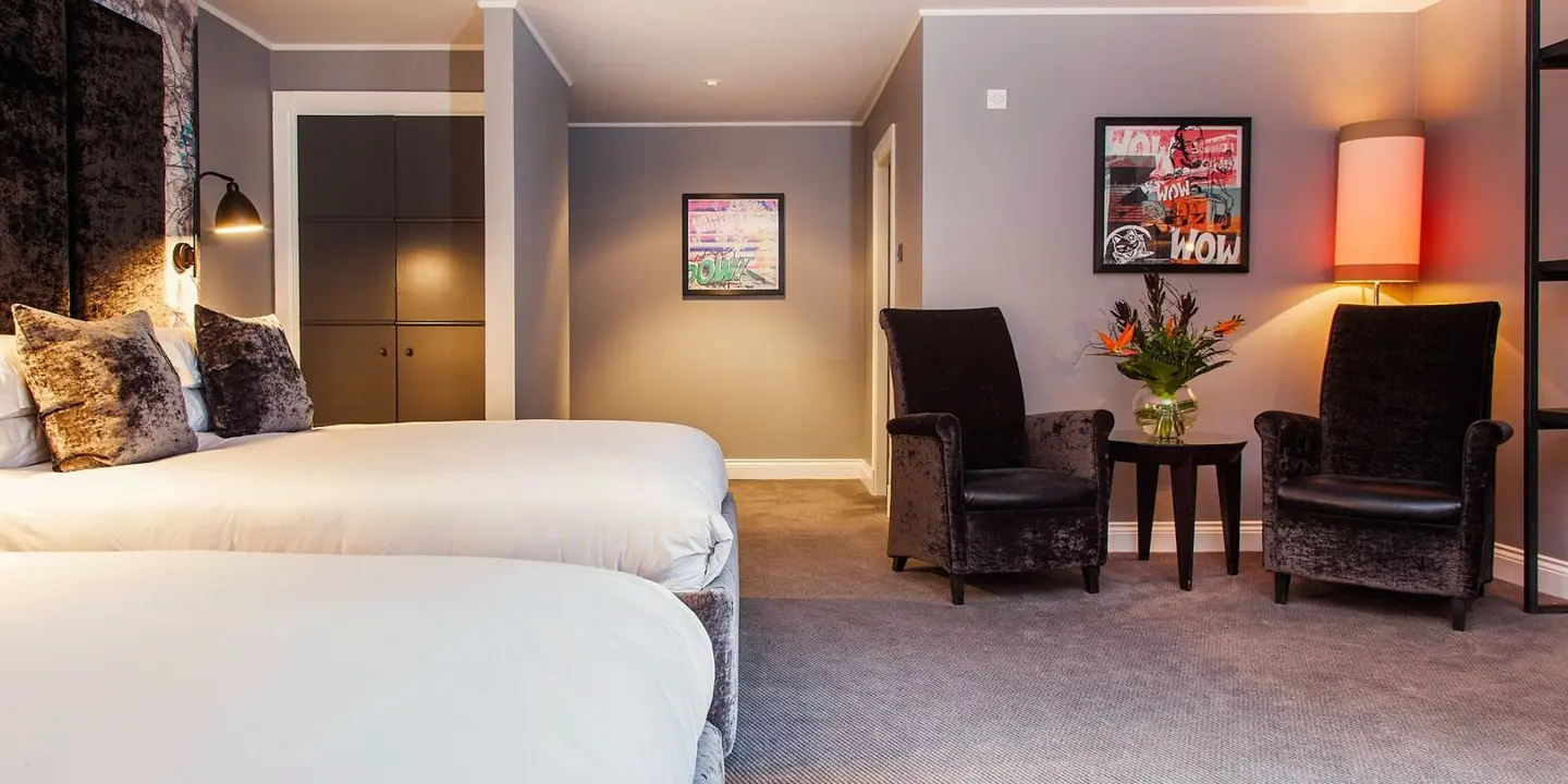 A hotel room with 2 grey velvet beds alongside 2 chairs and small table.