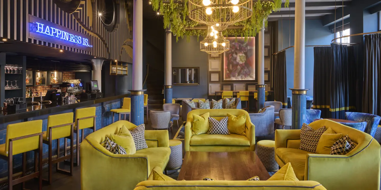 Yellow fabric sofas and bar stools prominently feature around a wooden table, with the bar in the background and a blue neon sign reading "happiness is" is displayed above the bar.