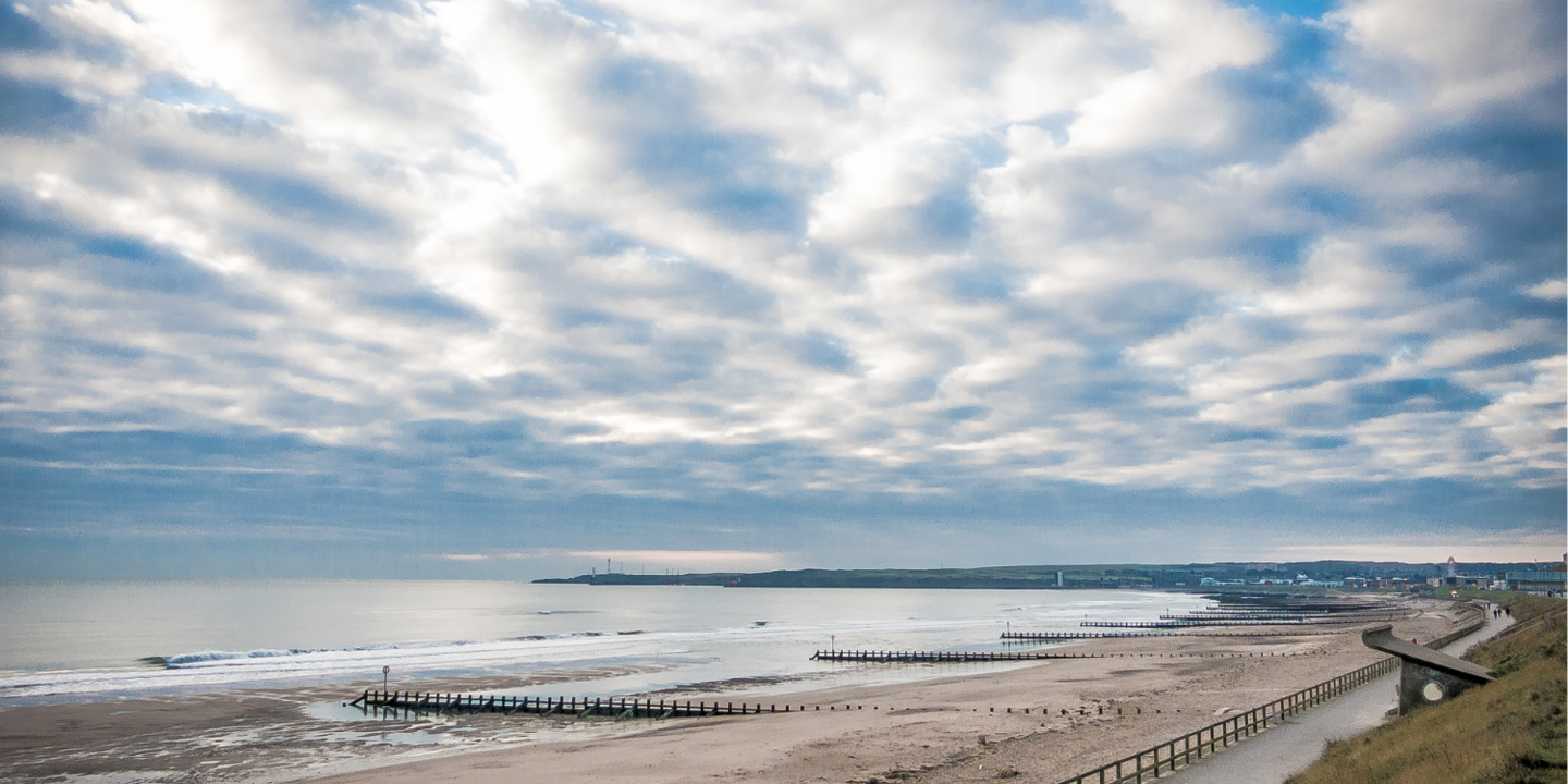 A wide angle picture of a sandy beach in Aberdeen. The sky is bright but overcast. A grassy dune is to the right of the image.