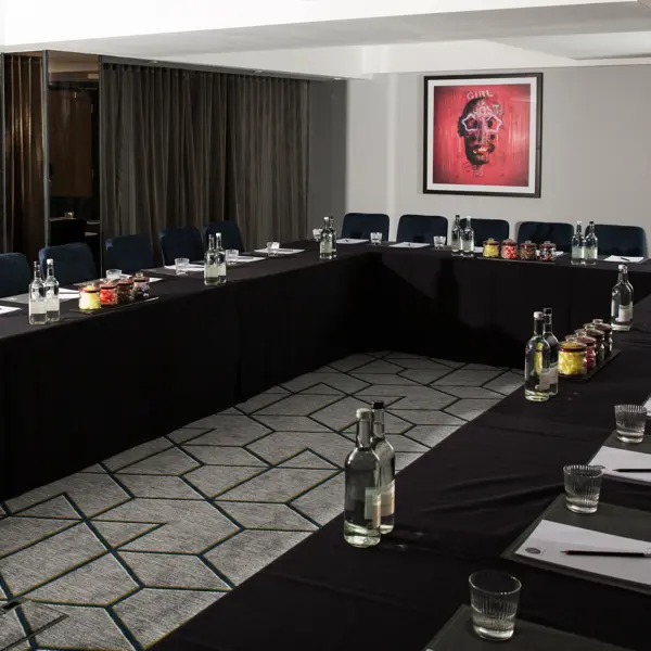 View of 3 tables arranged in a U shape with black table cloth and seats. Bottles of water, drinking glasses, writing pads and pencils, and jars of boiled sweets are on each table.