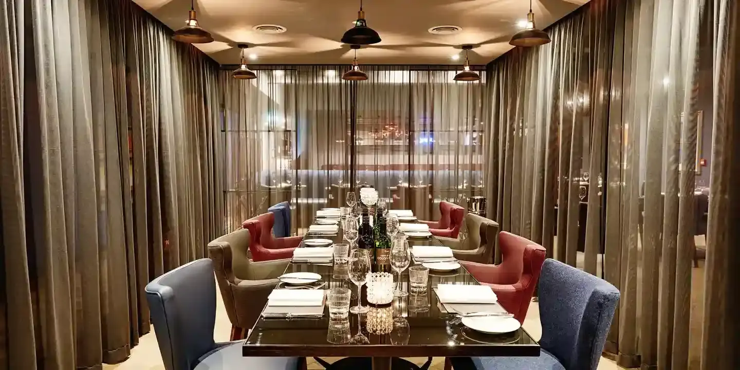 Another angle of The Cage dining room featuring a lengthy table adorned with blue, red and gold chairs, curtained glass walls, and brass light fixtures.