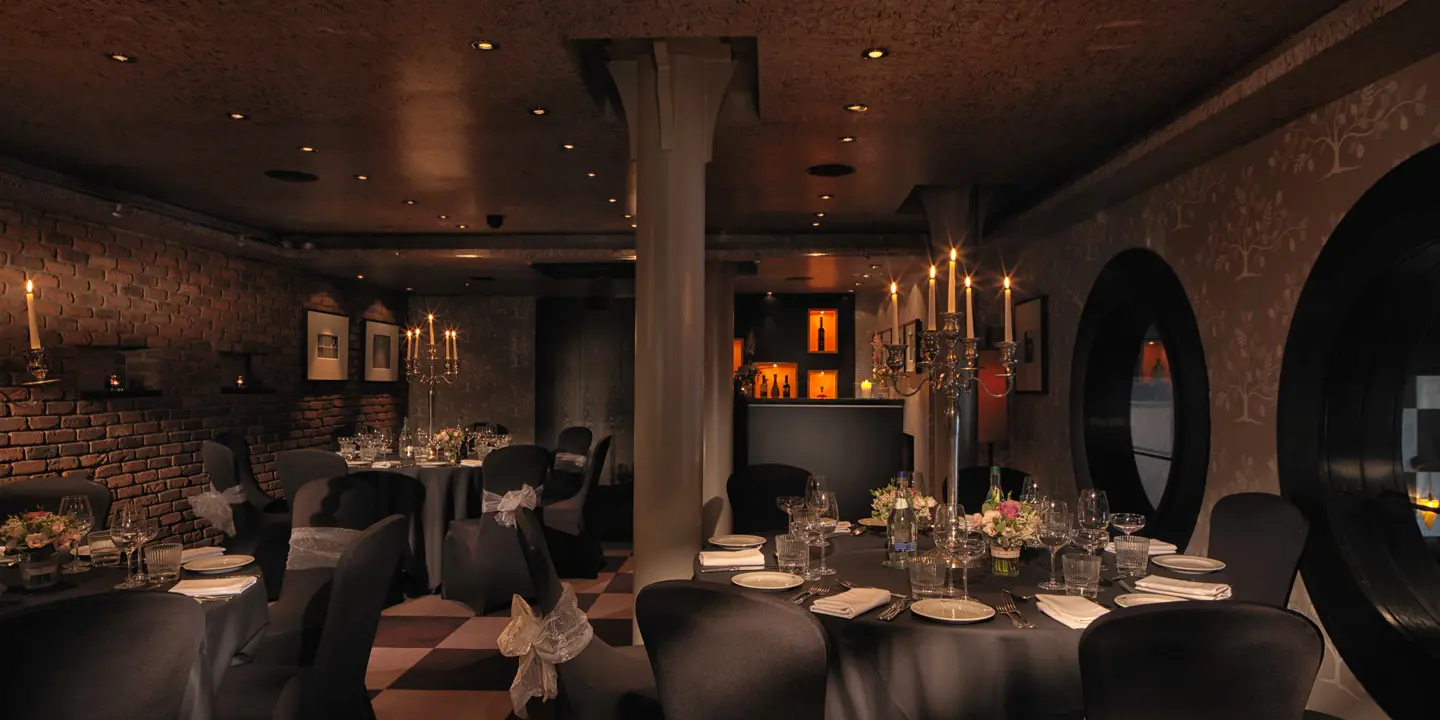 Private dining room set up for a wedding or similar event. Chairs featuring grey covers and white ribbon. Tall silver candelabra on each of the two round tables in view. 