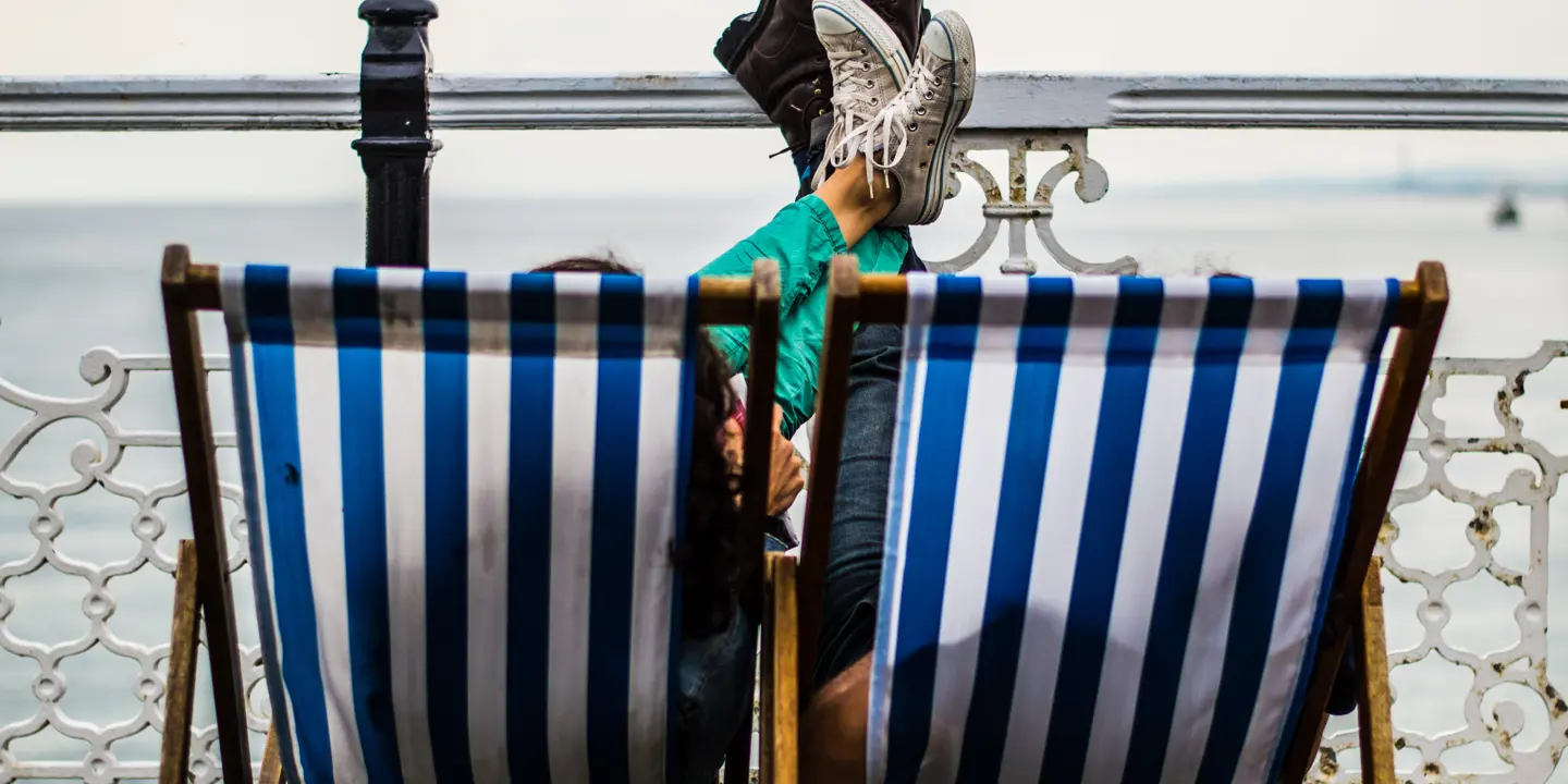 Two individuals seated on blue and white striped deck chairs, with feet resting on beachfront railing.