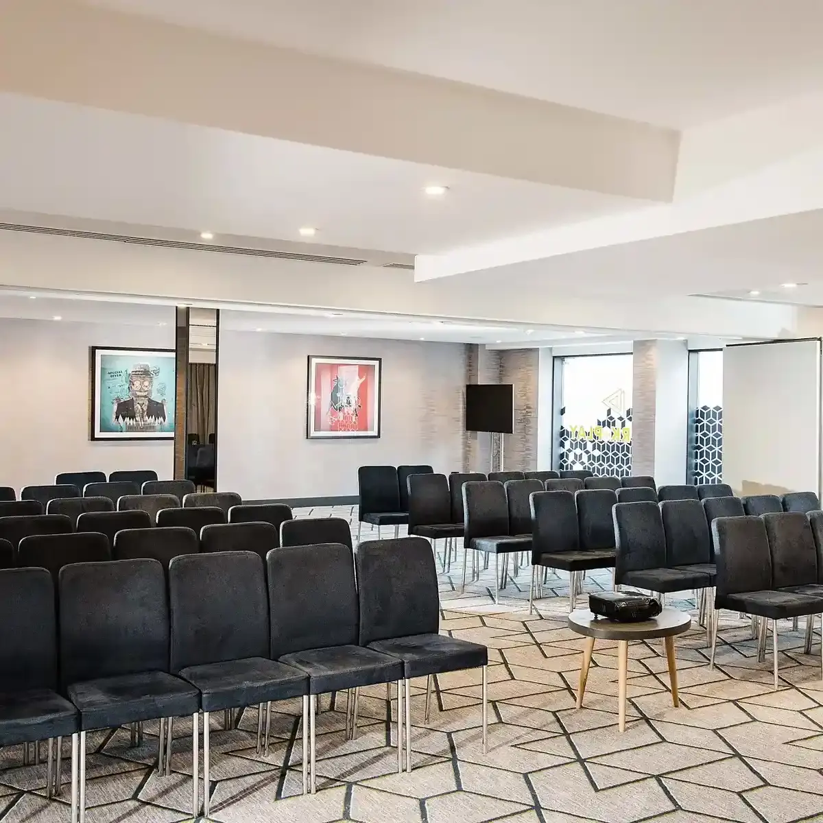 A meeting room set up for a seminar with rows of black fabric and metal seats, a geometric patterned cream and grey carpet, and a projector.