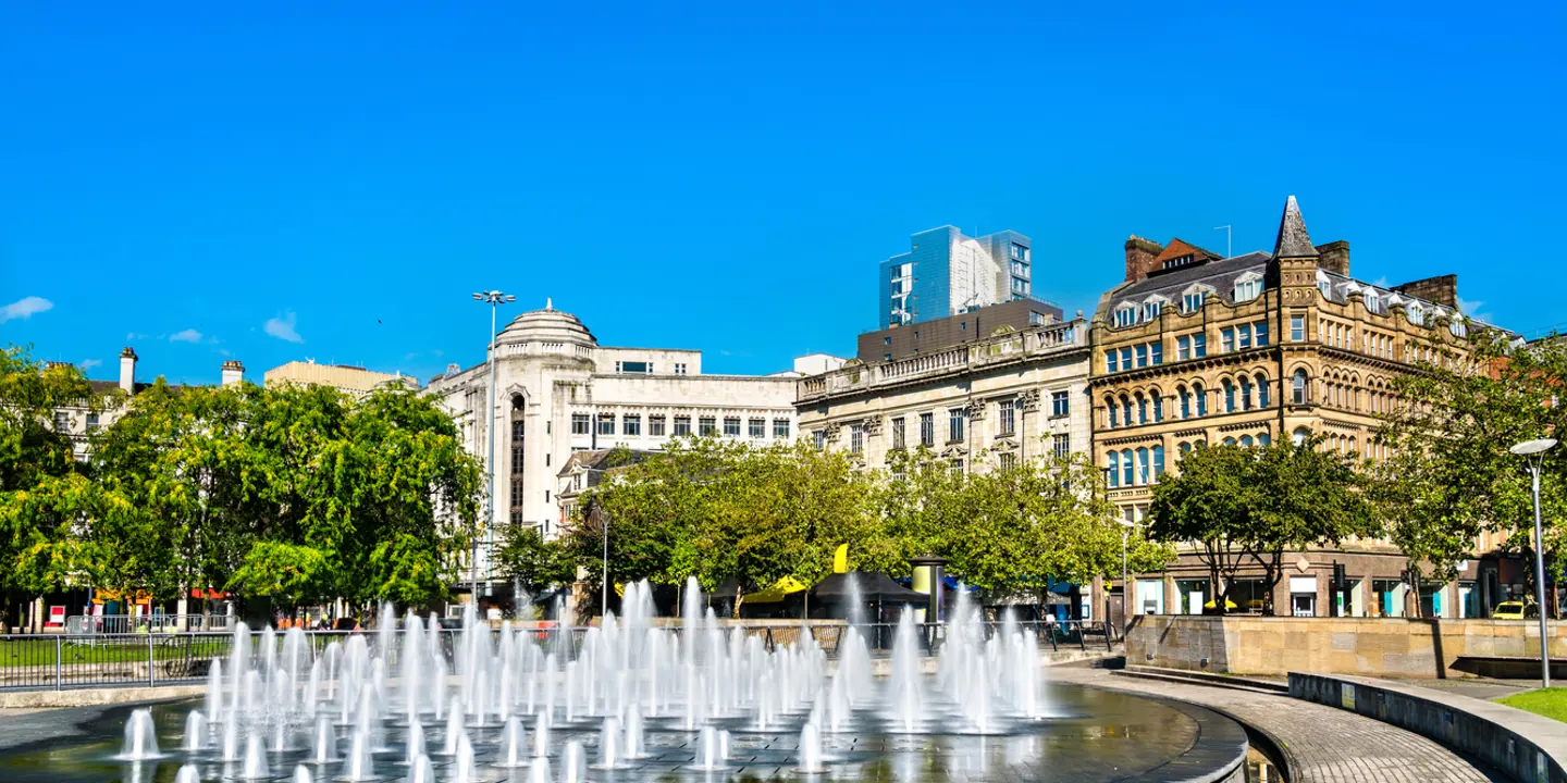 A fountain situated in a park with buildings visible in the background.